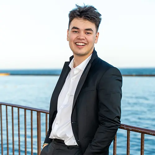 James Deogrades in a black suit, standing in front of a blue lake, smiling at the camera.